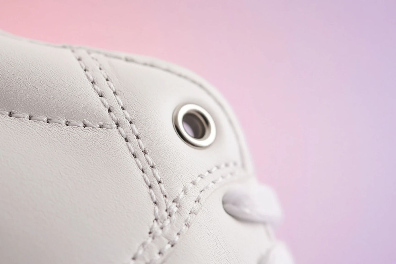 Macro crop of a white leather sneaker's stitched eyelet against a pink gradient